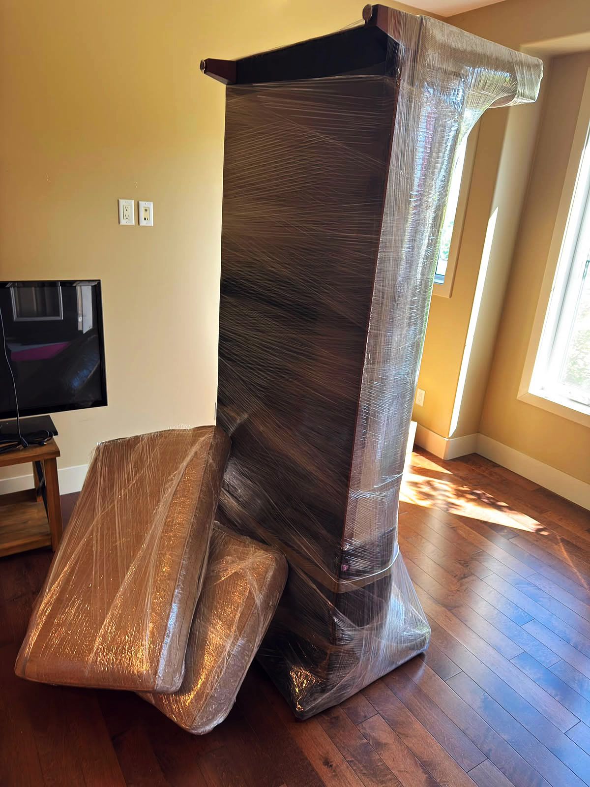 Tall wooden cabinet and seat cushions stretch-wrapped for a move in a sunlit living room